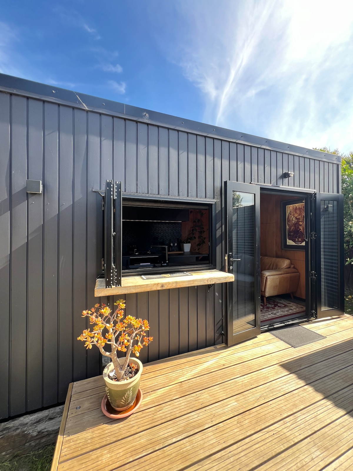 An exterior view of the bespoke tiny house highlights a sleek, dark grey façade with large, open windows. A small countertop is positioned beneath one window, adorned with a potted plant. The wooden deck provides a warm contrast to the contemporary design, complemented by the clear blue sky.
