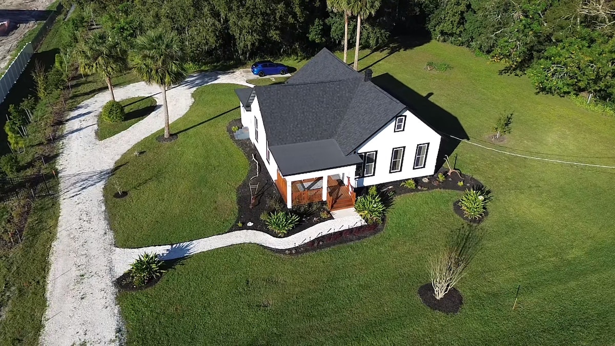 An aerial view captures The 1917 Farmhouse surrounded by lush greenery and well-kept landscaping. A winding pathway leads to the front porch, with a vibrant blue car visible in the driveway, enhancing the home's inviting exterior.