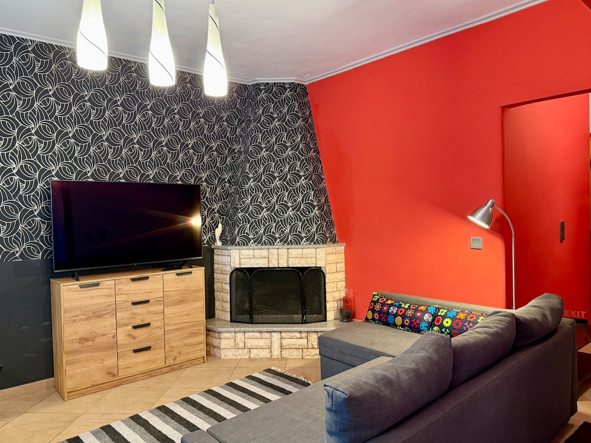 A modern living area features a large television mounted against a patterned wall. A cozy gray sofa is paired with a colorful cushion along the edge. A stone fireplace adds warmth alongside three hanging light fixtures, enhancing the inviting atmosphere of the room.