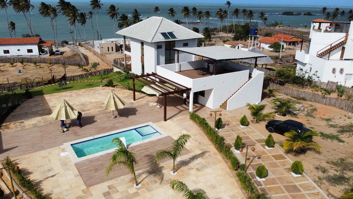 An aerial view captures the expansive outdoor area, featuring a swimming pool surrounded by lounge chairs and shaded umbrellas. Lush greenery and decorative plants enhance the space. The backdrop includes the tranquil ocean and palm trees, with neighboring structures visible in the distance.