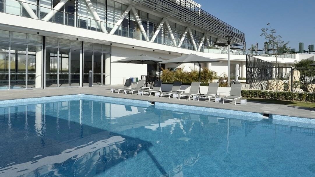 A recreational pool area is visible, featuring a clear blue water surface reflecting the surroundings. Several lounge chairs are arranged along the pool's edge. In the background, a modern building with large glass windows and shaded seating areas can be seen, surrounded by landscaped greenery.