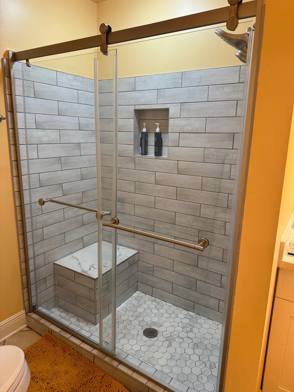 A modern shower stalls with glass sliding doors is featured, showcasing a tiled interior. A built-in shelf holds two bottles, while a marble bench provides seating. The floor is laid with white hexagonal tiles, and a yellow mat adds a touch of color in the foreground.