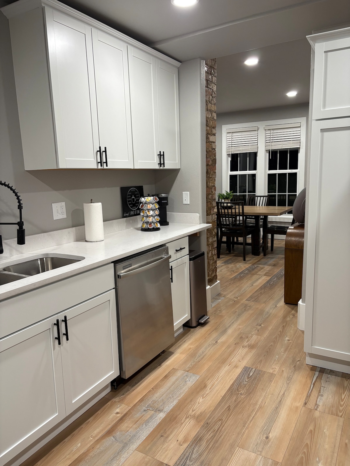 The kitchen features sleek white cabinetry and a modern stainless steel dishwasher. A deep sink is complemented by a stylish black faucet, while a coffee station is arranged on the countertop. A dining area is visible in the background, with a wooden table and four chairs.