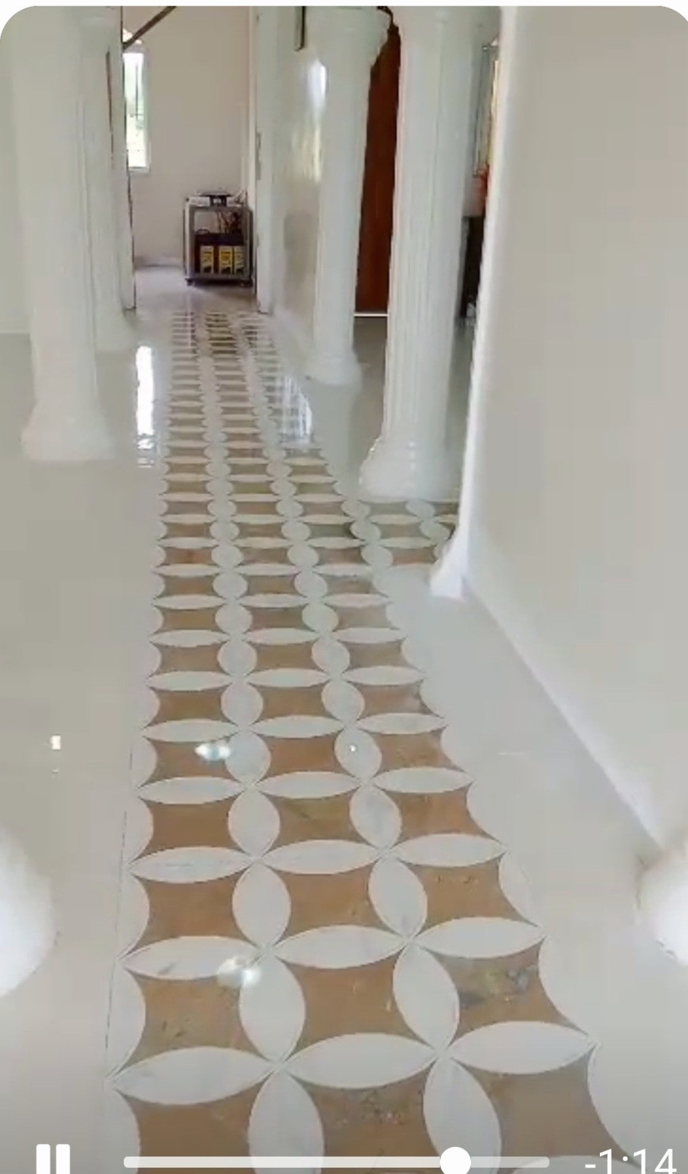 A clean, tiled hallway is highlighted with a geometric pattern featuring light and earthy tones. Tall columns frame the pathway, while ample natural light brightens the interior, creating an open and welcoming feel.