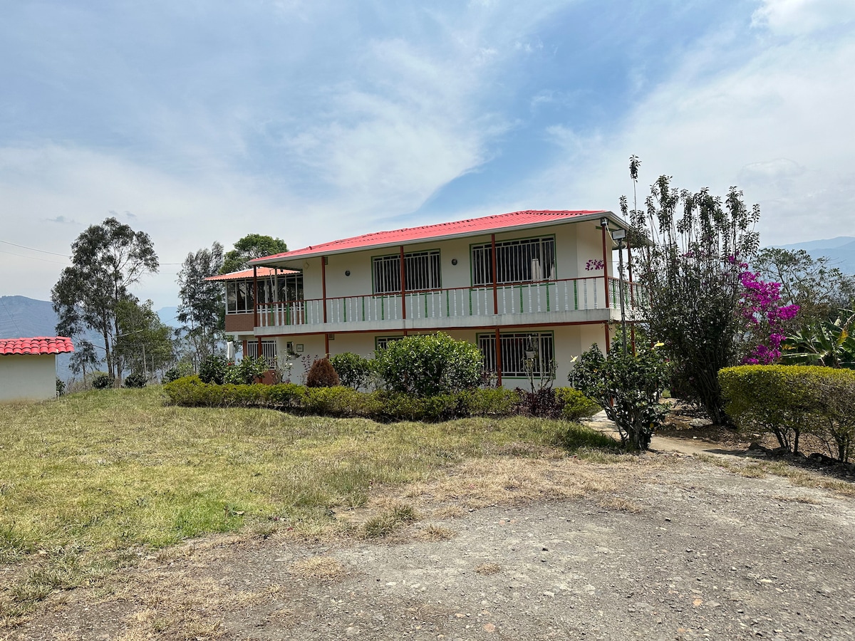 A two-story building is surrounded by lush greenery and flowering plants. The property features a red tiled roof and spacious balcony areas with white railings. A well-maintained lawn stretches in front, offering a serene outdoor setting. Mountains are seen in the background.