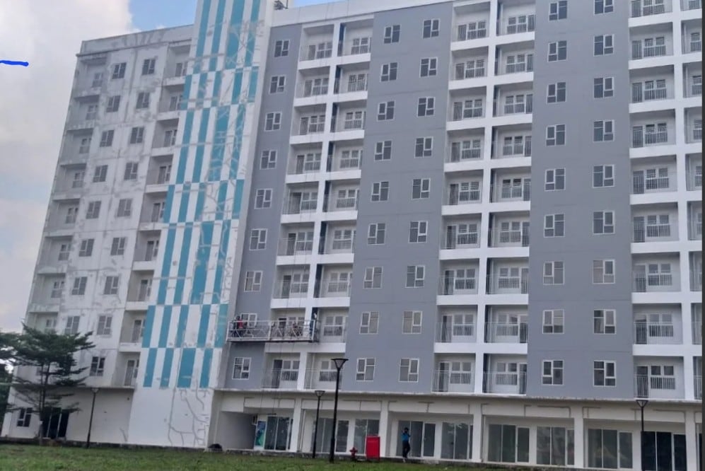 A multi-story building features a modern facade with alternating stripes of grey and blue. Numerous windows line the exterior, allowing natural light to fill the interiors. Surrounding grassy areas are visible, offering a space for relaxation and outdoor activities.