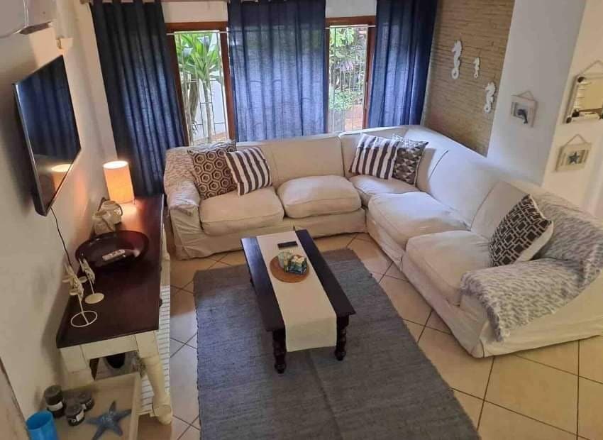 A spacious lounge area features a white sectional sofa with decorative throw pillows, arranged around a dark coffee table. Soft lighting from a lamp complements the natural light filtering through partially open curtains. Decorative wall elements enhance the relaxed atmosphere.