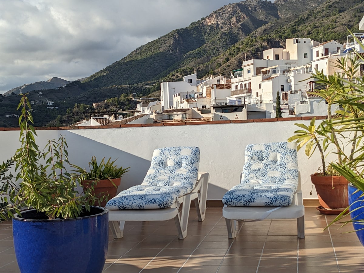 A spacious rooftop terrace features two sun loungers with blue cushioned fabric. Green plants are positioned in terracotta pots, providing a natural touch against the backdrop of rolling hills and whitewashed buildings in the distance.