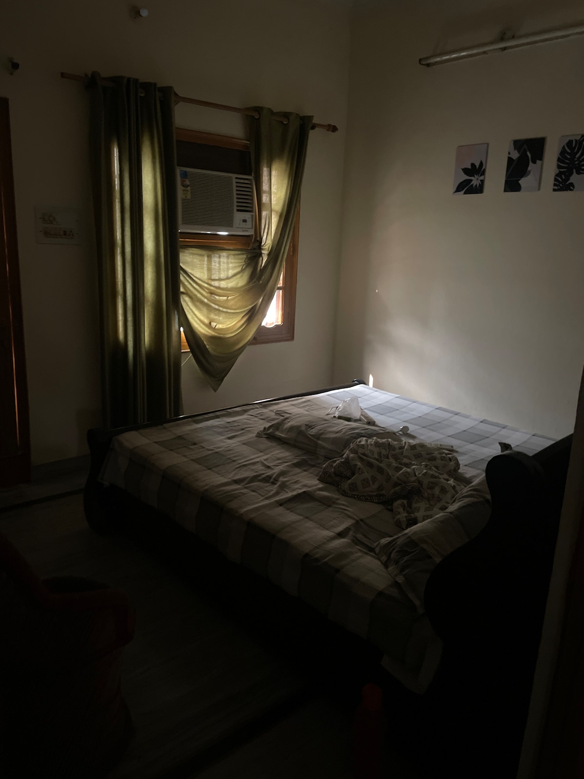 A serene bedroom is viewed, featuring a spacious bed with a checkered comforter. Soft natural light enters through a window adorned with flowing green curtains. Above the bed, three framed art pieces provide a subtle touch to the calming atmosphere.