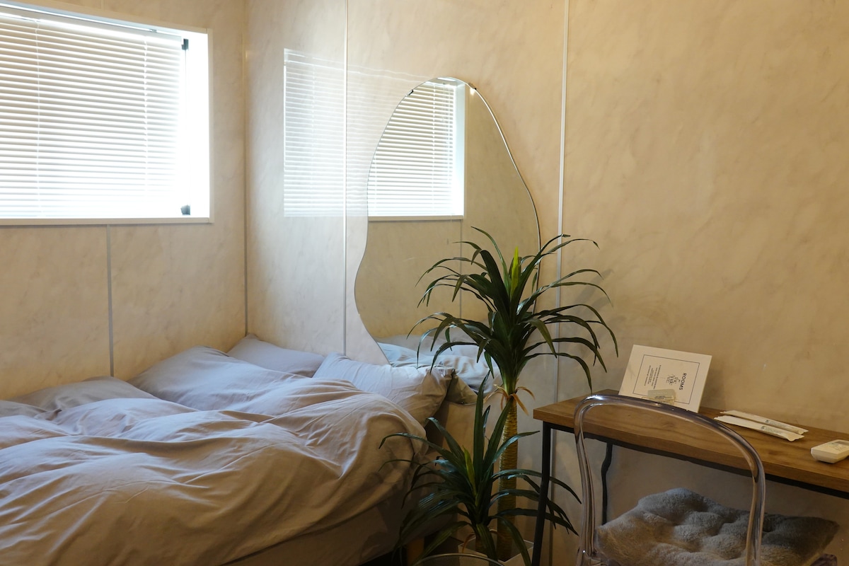 A cozy room features a spacious bed with neutral bedding, complemented by a decorative plant beside it. A large mirror reflects natural light from the nearby window, and a wooden table with minimalist decor completes the inviting atmosphere.
