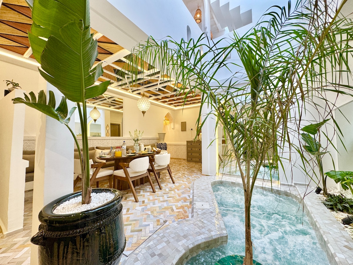 An indoor space features a cozy dining area with a large table set for meals. Sunlight filters through the open ceiling, highlighting the lush green plants beside a bubbling pool. Textured tiles provide a warm aesthetic throughout the area.