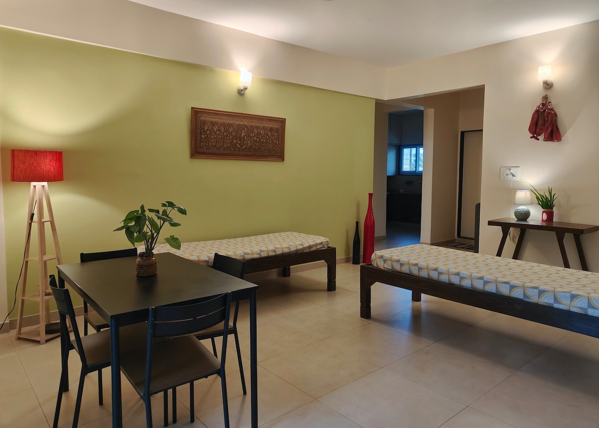 The living area features two single beds with decorative bedding against a green wall. A small dining table with four chairs occupies the center space, while a potted plant and two decorative vases add subtle accents. Soft lighting contributes to a relaxed atmosphere.