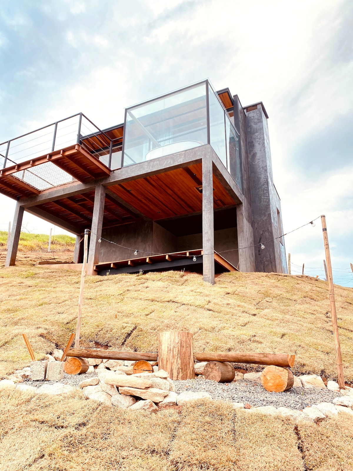 A modern house is set against a hillside, showcasing a blend of glass and concrete in its design. The structure features large windows and a spacious deck, with natural landscape elements such as a stone pathway and logs visible at the base.