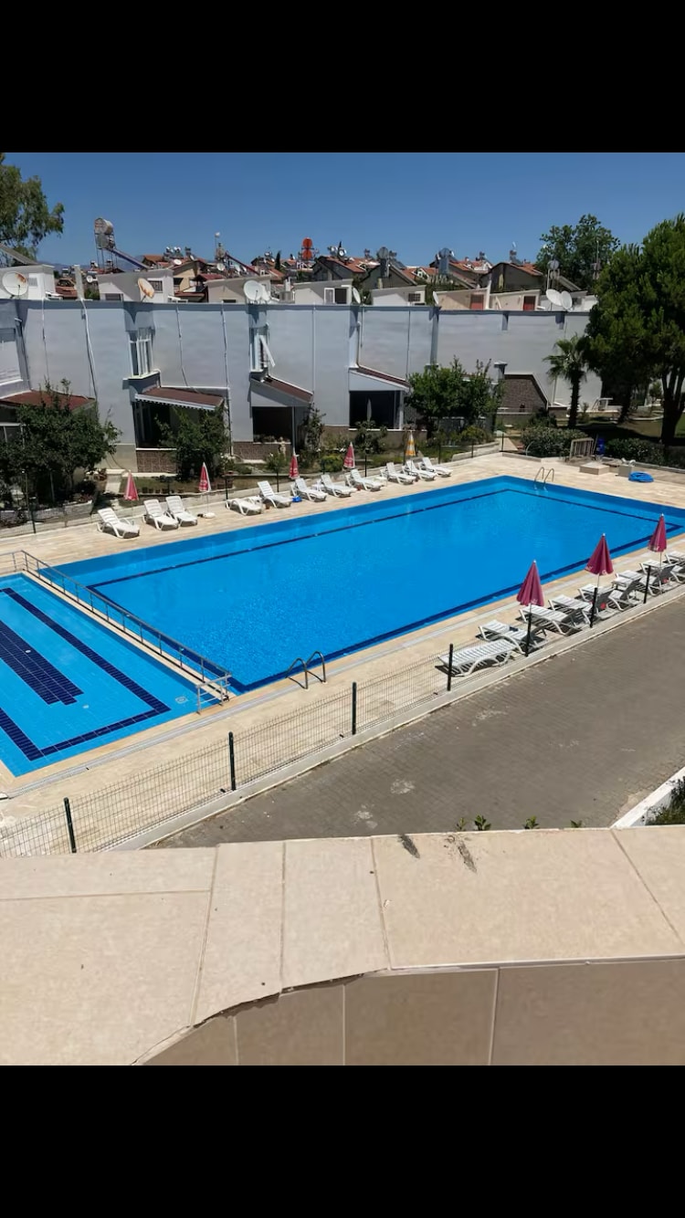A large outdoor swimming pool is surrounded by lounge chairs. A few umbrellas provide shade alongside the pool. The area is bordered by a pathway, and trees can be seen in the background, with buildings visible in the further distance.