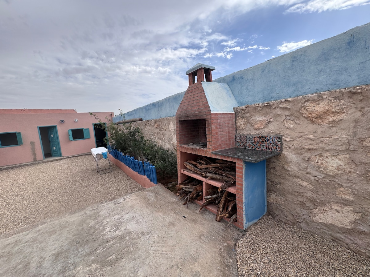 An outdoor area features a traditional brick barbecue, complete with a chimney and neatly stacked firewood. Surrounding the barbecue is a stone wall and a gravel surface, creating a rustic setting. Nearby, a table can be seen against a colorful backdrop of blue and pink walls.
