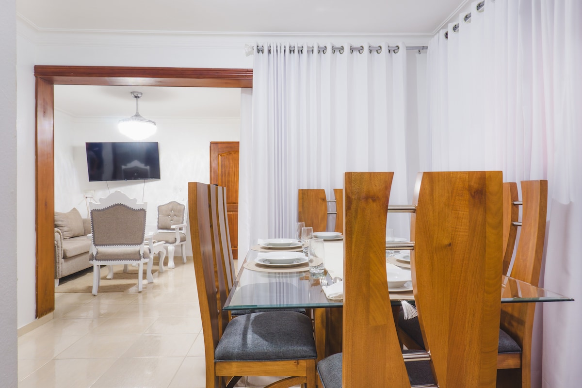 A dining area features a glass table surrounded by tall wooden chairs. Formal table settings are arranged neatly. White curtains frame the space, providing a soft contrast to the warm wood tones. In the background, a comfortable seating area is visible, enhancing the inviting layout.