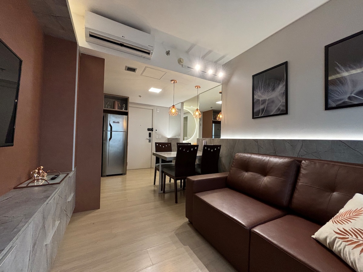 A modern living space is depicted, featuring a brown leather sofa and a dining table with four chairs. An air-conditioning unit and a stainless steel refrigerator enhance the functionality of the area. Decorative wall art adds a subtle touch to the inviting atmosphere.