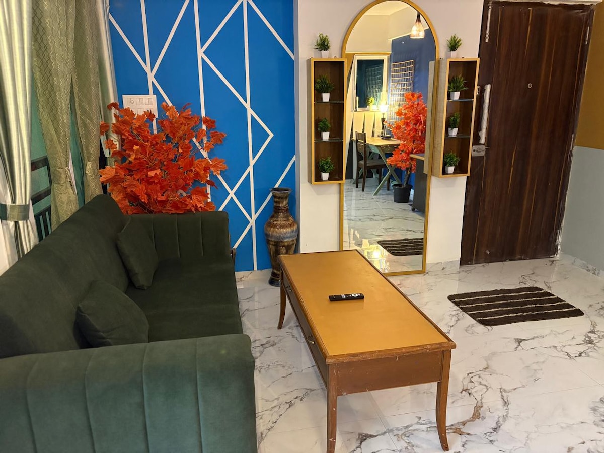 A contemporary living area features a green sofa facing a sleek coffee table. A full-length mirror is positioned against a blue geometric wall, complemented by a decorative arrangement of orange leaves. Light marble flooring enhances the spacious ambience, with a doorway visible in the background.