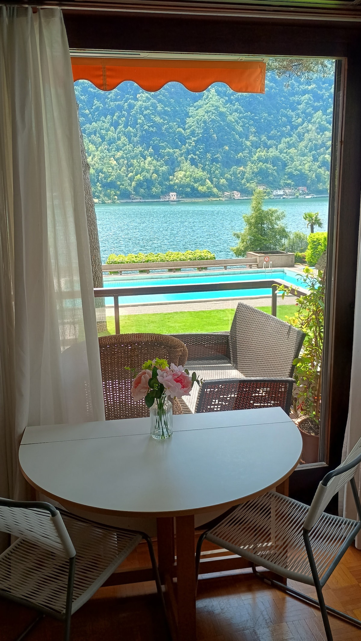 A table is set by the window, featuring a small vase with fresh flowers. Outside, a picturesque view of the lake and lush mountains can be seen. Two wicker chairs are positioned on the balcony, inviting relaxation while enjoying the surrounding nature.