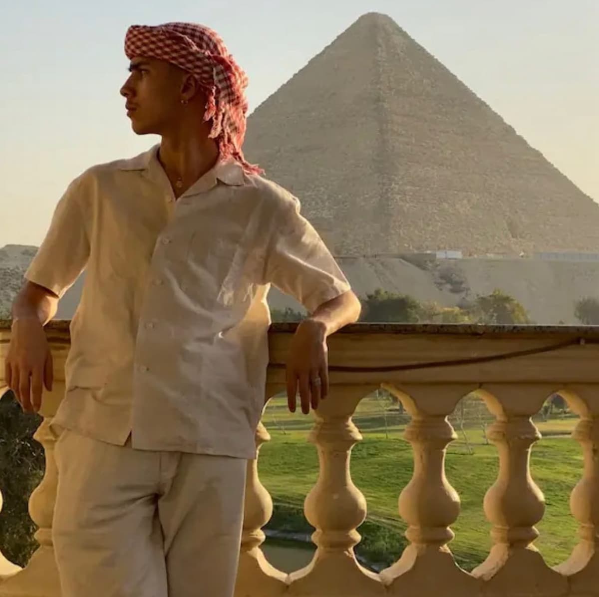 A person stands on a balcony, looking out towards the Giza Pyramid in the background. The balcony features a decorative railing and offers a clear view of the pyramid, surrounded by green grass and trees.
