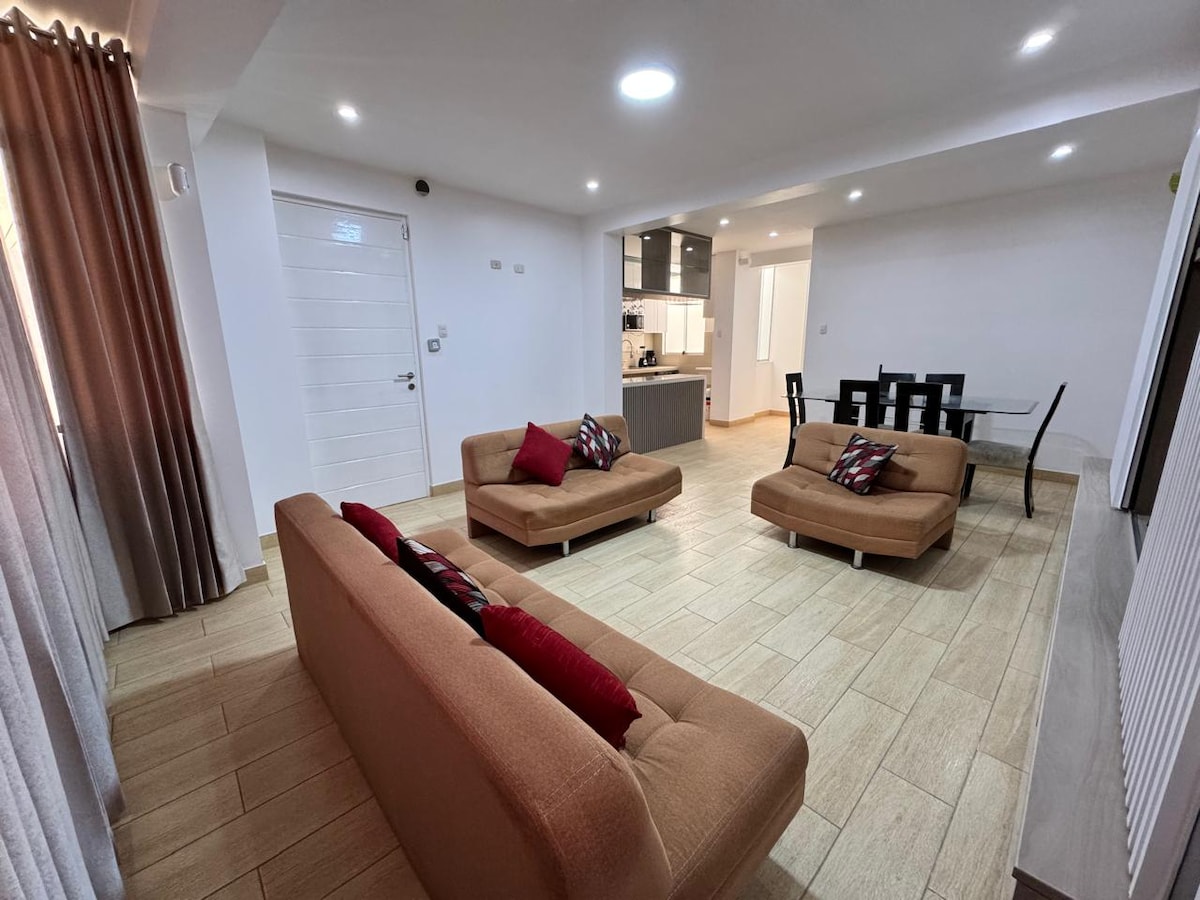 A spacious living area is presented, featuring two cozy sofas with decorative cushions. A dining area is visible in the background, accompanied by a modern kitchenette. Natural light filters through the large windows, enhancing the inviting ambiance of the room.