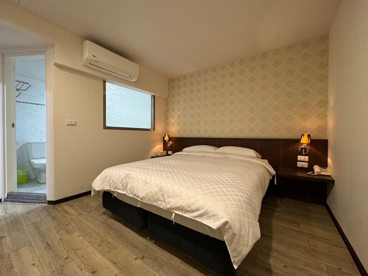 A spacious sleeping area features a large bed with a textured white duvet. Two bedside lamps provide soft lighting on each side of the bed. A window allows natural light to enter, and a minimalist wooden floor complements the room's design.