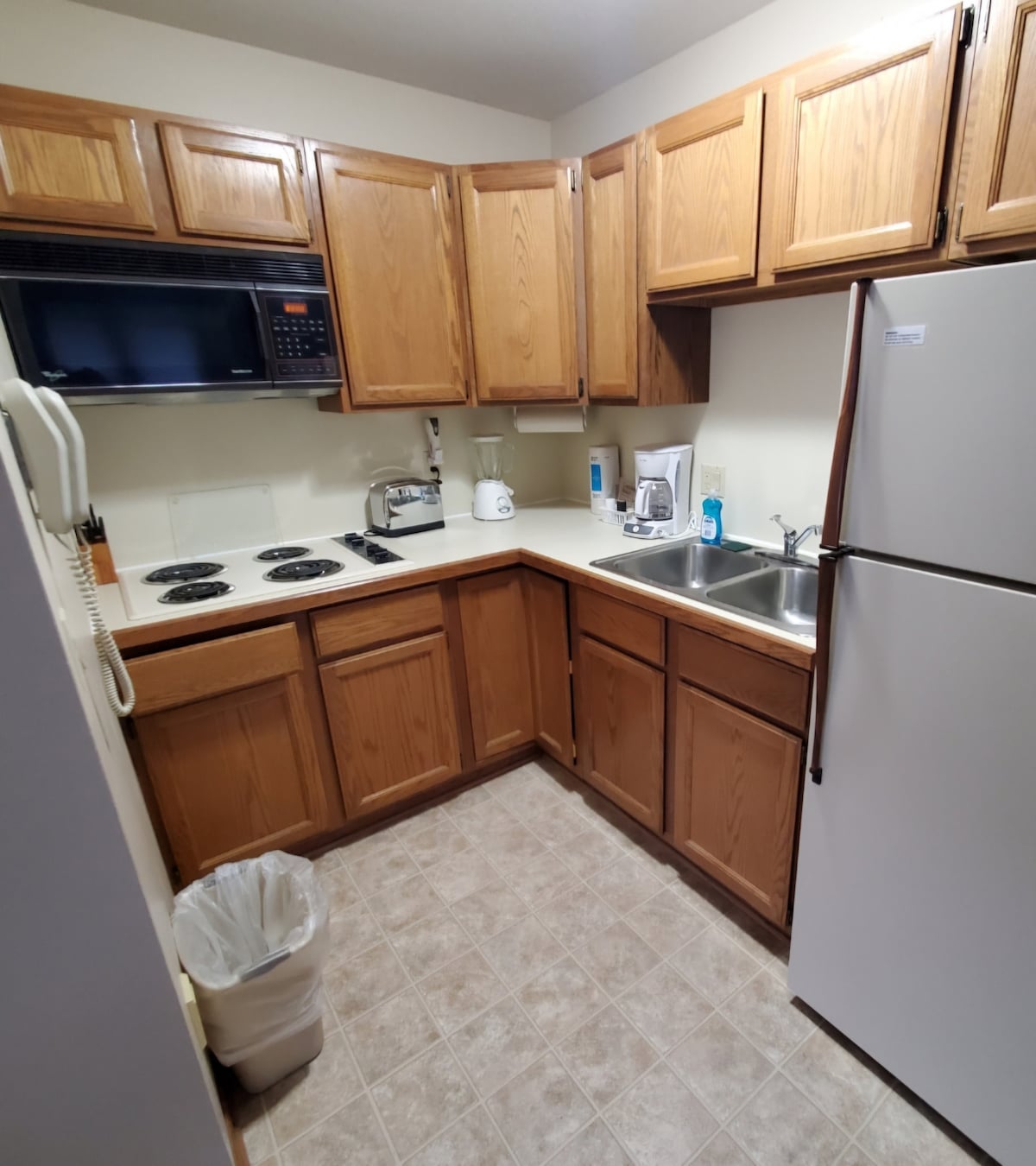 The kitchen area features wooden cabinets with a microwave and a stove with two burners. Stainless steel appliances, including a refrigerator and a sink, are positioned next to each other. A trash can and various kitchen essentials are visible on the countertops.