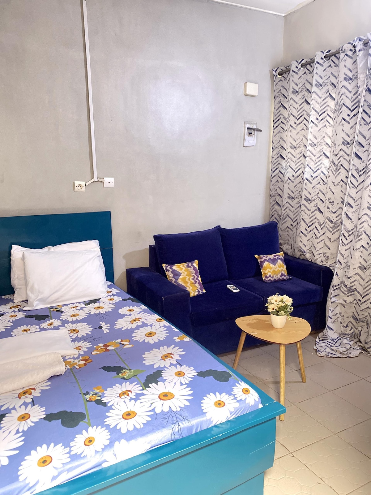A cozy room features a bed with a floral-patterned bedspread and a blue velvet sofa with decorative pillows. A small round table sits in front of the sofa, with a floral arrangement adding a touch of color. Light-colored curtains frame the window.