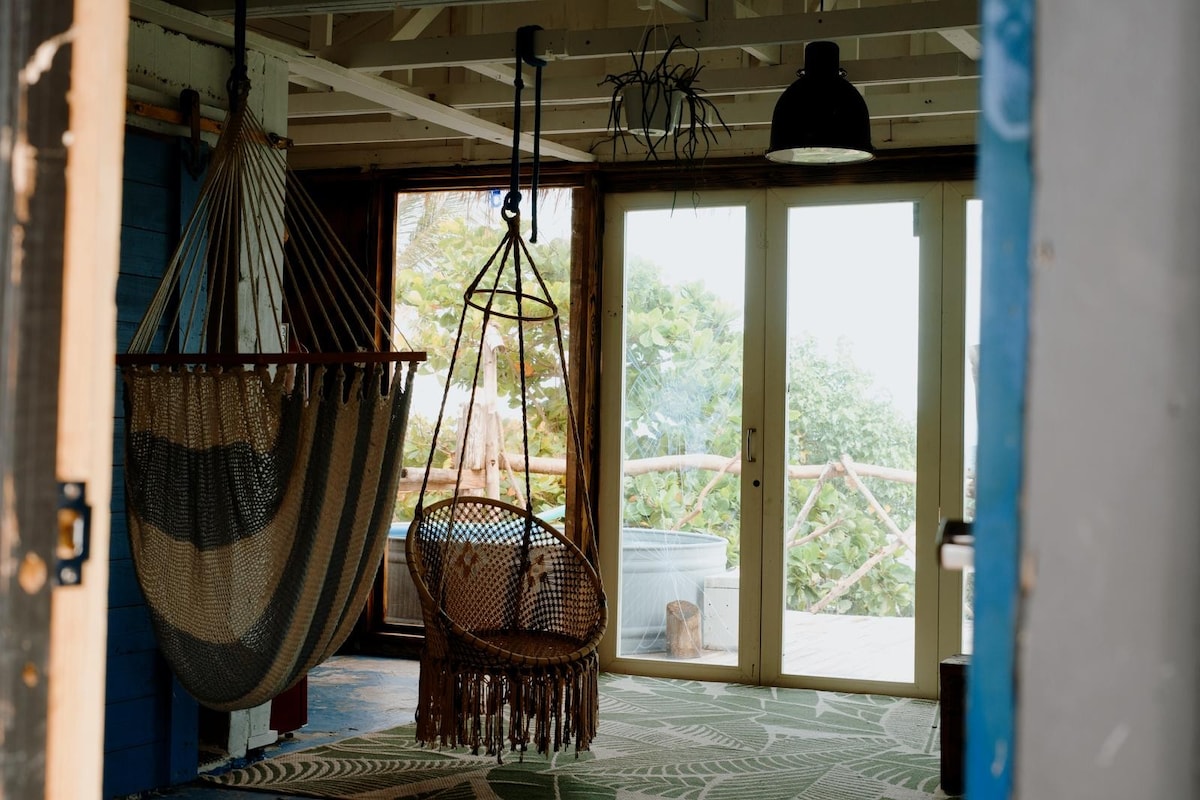 A bright interior space features two hanging chairs, one with a striped design and the other intricately woven. Large glass doors lead to a view of greenery outside, while a patterned rug adds texture to the floor. Natural light filters through the windows.