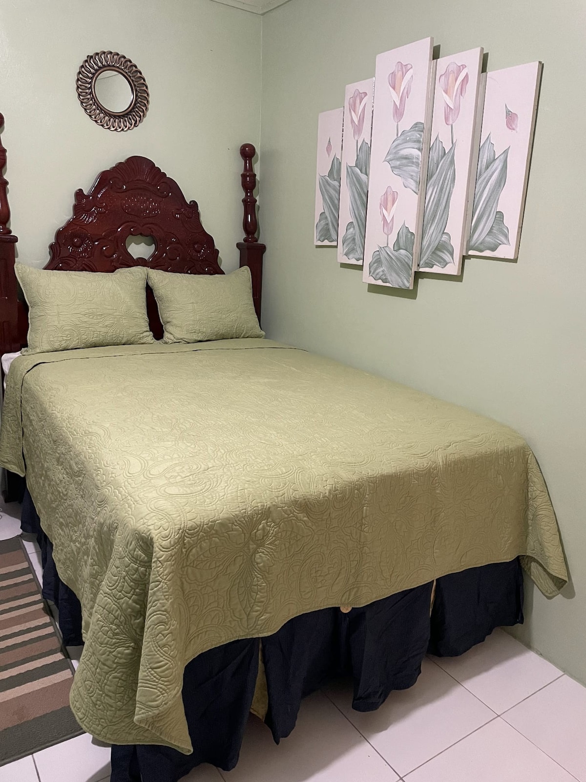 A well-appointed sleeping area features a neatly made bed with a light green quilt and matching pillows. Above the bed, a series of floral wall art adds a touch of decor. A round mirror complements the warm tones of the furnishings.
