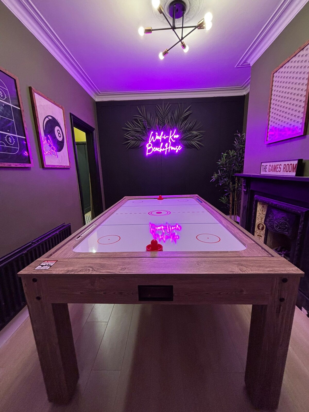 A games room is highlighted by a wooden air hockey table positioned in the center. The walls feature dark paint, adorned with framed artwork and a neon sign stating 'The Games Room.' A decorative plant and a vintage fireplace add character to the space.