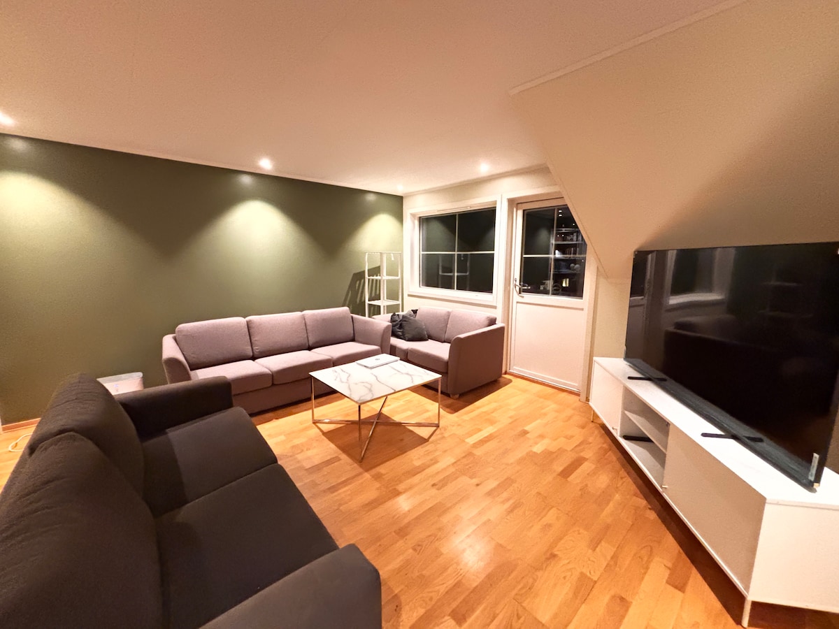 A spacious living room is featured, showcasing multiple seating arrangements with soft gray sofas surrounding a marble coffee table. Natural light enters through large windows, and a flat-screen television is positioned against a light-colored entertainment unit. Warm wooden flooring adds to the inviting ambiance.