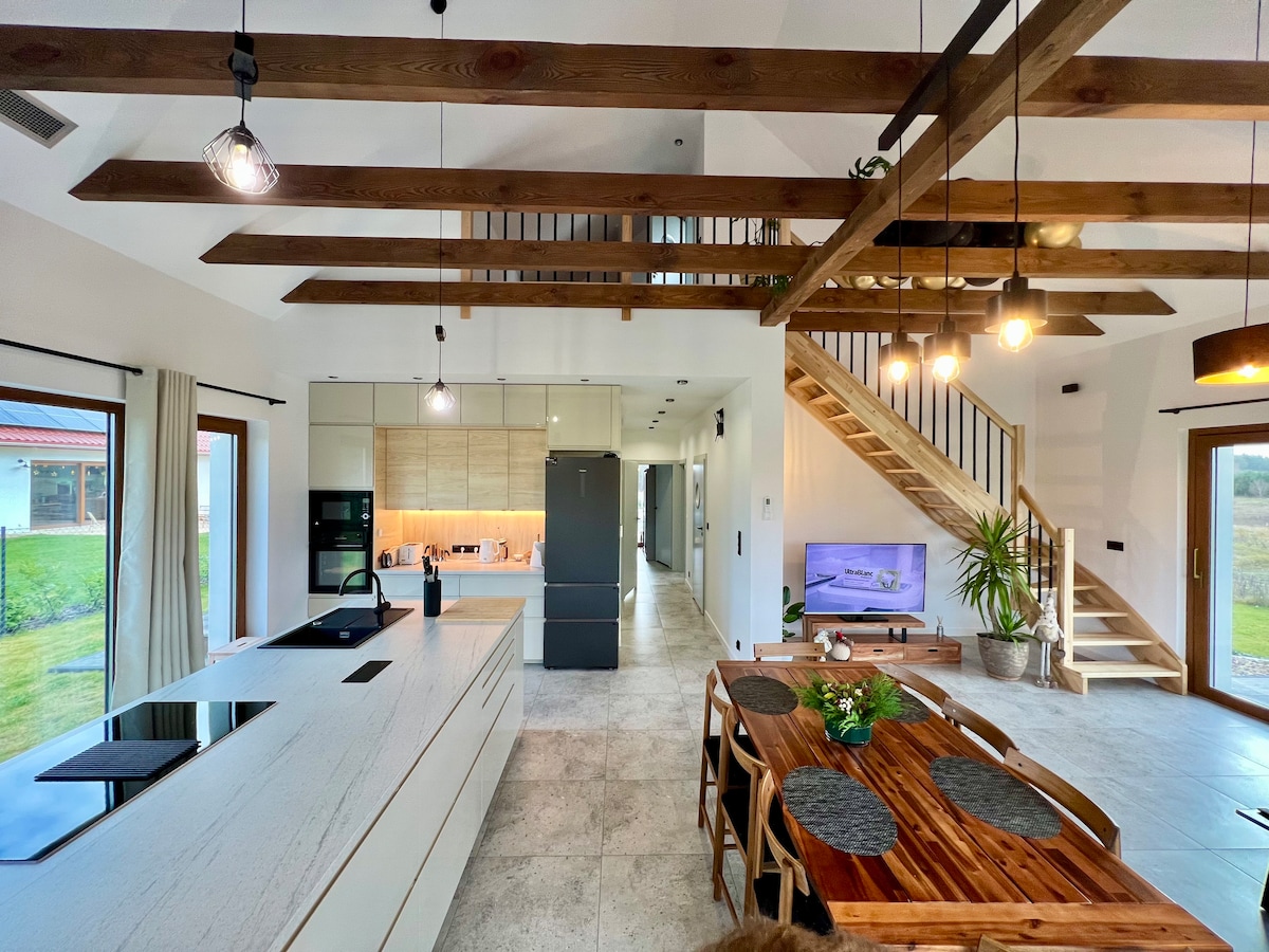 An airy open plan living area showcases a modern kitchen with sleek cabinetry and an island for meal preparation. A wooden dining table sits invitingly beside large windows, while stairs lead to the upper level. Natural light floods the space through various windows.