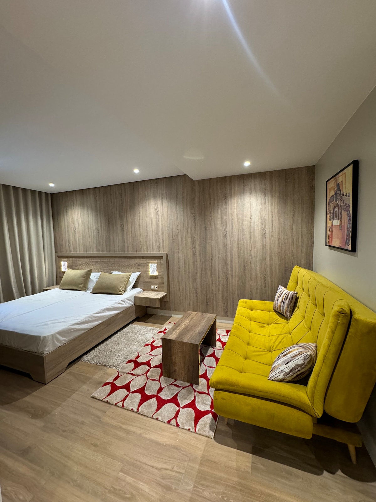 A cozy sleeping area features a spacious bed with neutral bedding, complemented by two decorative pillows. A bright yellow sofa adds a pop of color, while a wooden coffee table sits centrally on a patterned area rug. Soft lighting illuminates the warm wood-tone walls.