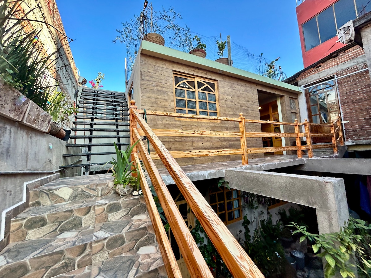 A charming exterior showcases a wooden deck leading to the entrance of the apartment, surrounded by lush greenery. A stone pathway guides visitors up to the entrance, while stairs ascend to a rooftop garden adorned with plants, enhancing the inviting atmosphere of the space.