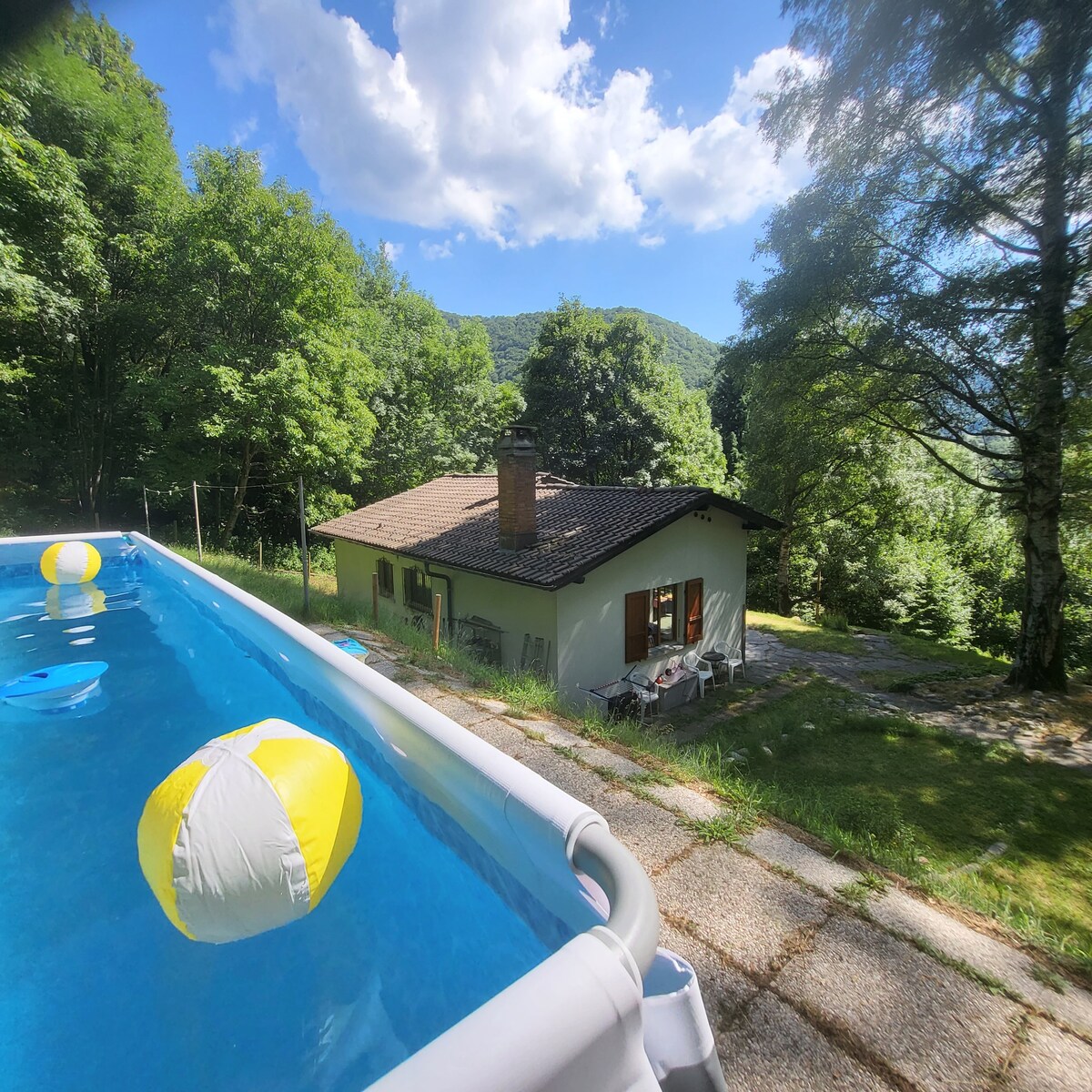 A serene outdoor view of the house set against a backdrop of lush greenery and mountains. A swimming pool with colorful beach balls is visible in the foreground, while the house, featuring a sloped roof and patio area, sits harmoniously within the landscape.