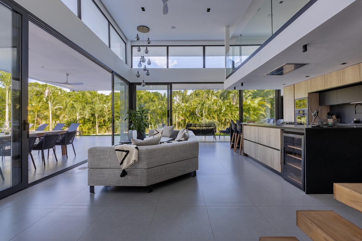 The open-plan living area is characterized by expansive glass walls that invite natural light, connecting indoor and outdoor spaces. A comfortable sofa is positioned centrally, complemented by a well-equipped kitchen featuring a large island. Tropical views are visible through the glass, enhancing the serene environment.