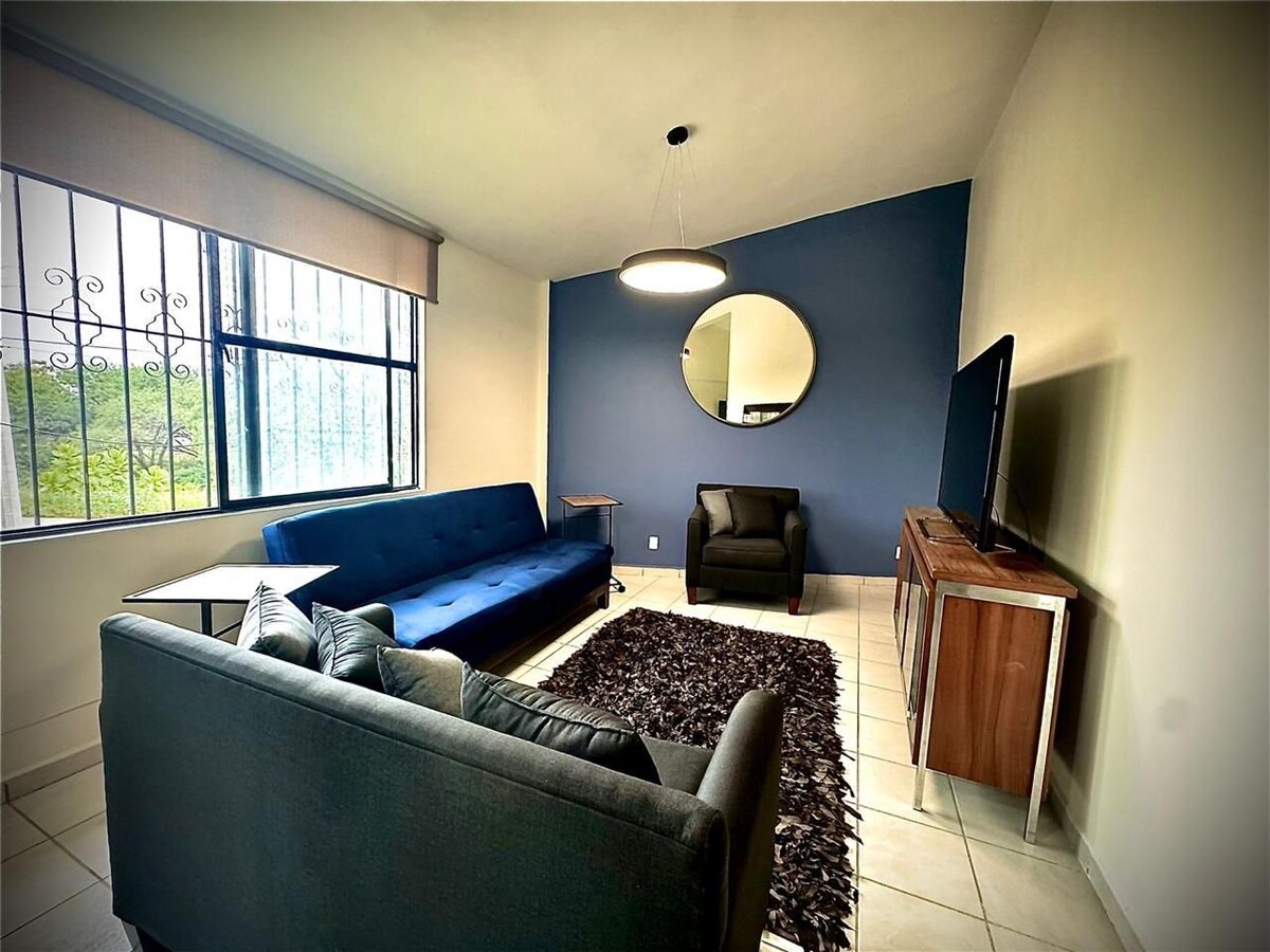 A modern living area features a blue sofa bed and a dark armchair positioned against a neutral wall. A large round mirror reflects light, and a flat-screen television sits on an accent table. Natural light filters in through the window adorned with blinds.