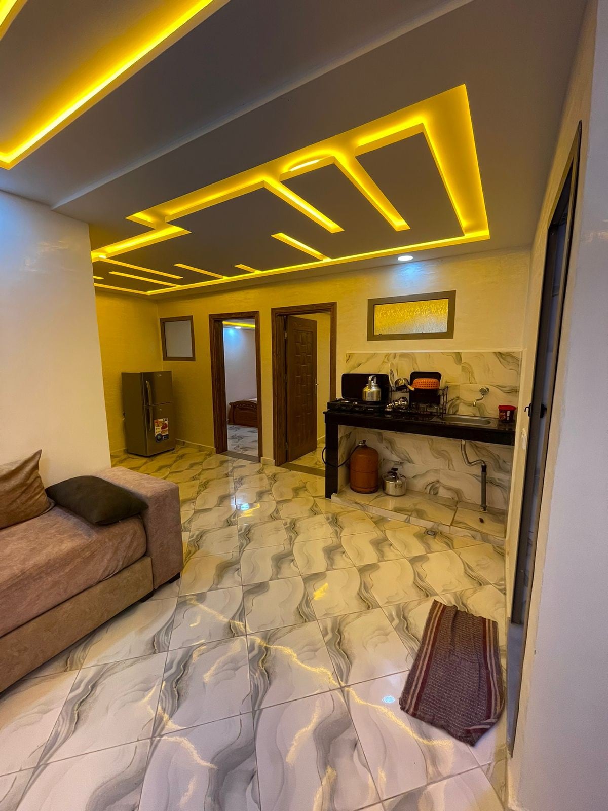A modern living area features soft seating in a neutral tone. Bright LED ceiling lights illuminate the space, accentuating the glossy patterned tile floor. Open doorways lead to additional rooms, while a small kitchenette is equipped with essential appliances.