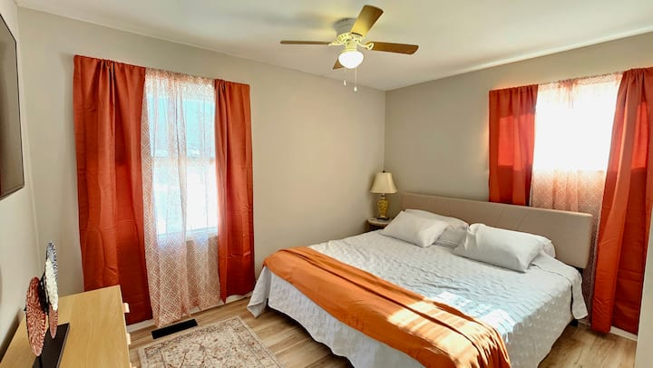 Manželská posteľ, komoda, nočný stolík, lampa, zatemňovacie závesy v miestnosti, televízor a stropný ventilátor.