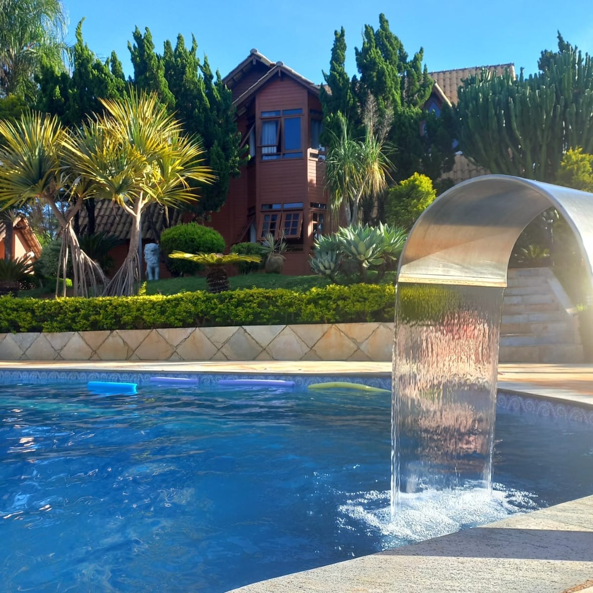 A serene outdoor pool is featured, with a flowing water feature creating a gentle cascade. Lush greenery surrounds the area, and a rustic building is visible in the background, enhancing the natural setting.