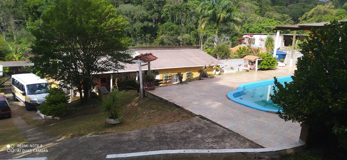 A spacious outdoor area is shown featuring a swimming pool surrounded by a tiled patio. Lush greenery decorates the landscape, with trees and plants providing a natural setting. The main building is visible, showcasing a covered porch with seating. Additional structures are present in the background.