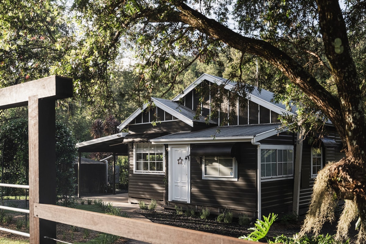 The cottage exterior is framed by lush greenery, showcasing large windows that allow natural light to fill the space. A simple fence runs along the property, complementing the rustic charm of the building with its dark wood siding and a welcoming front door.