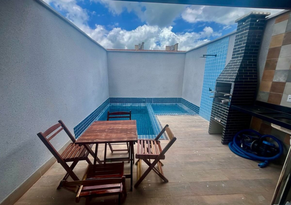 An outdoor space features a private pool surrounded by blue-tiled walls, offering a refreshing retreat. A small wooden table and four folding chairs are positioned nearby, alongside a built-in barbecue area adorned with a stone chimney.