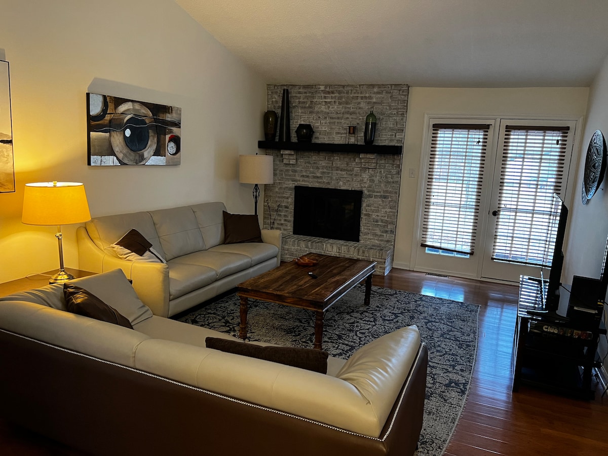The spacious living room features a combination of leather couches positioned around a wooden coffee table. A cozy rug lies beneath them. A fireplace is set into a brick wall, and large glass doors lead to an outdoor deck, allowing natural light to enter the space.