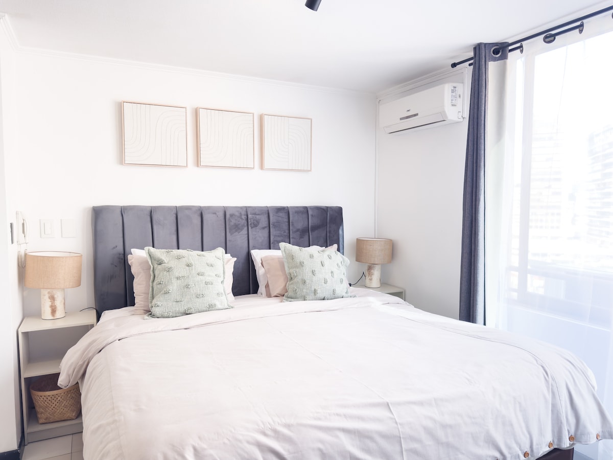 A bright bedroom features a spacious bed with soft linens, accented by two textured pillows. Three abstract art pieces are displayed above the headboard. Natural light filters through a sheer curtain, while a pair of bedside lamps and a small basket complete the setting.