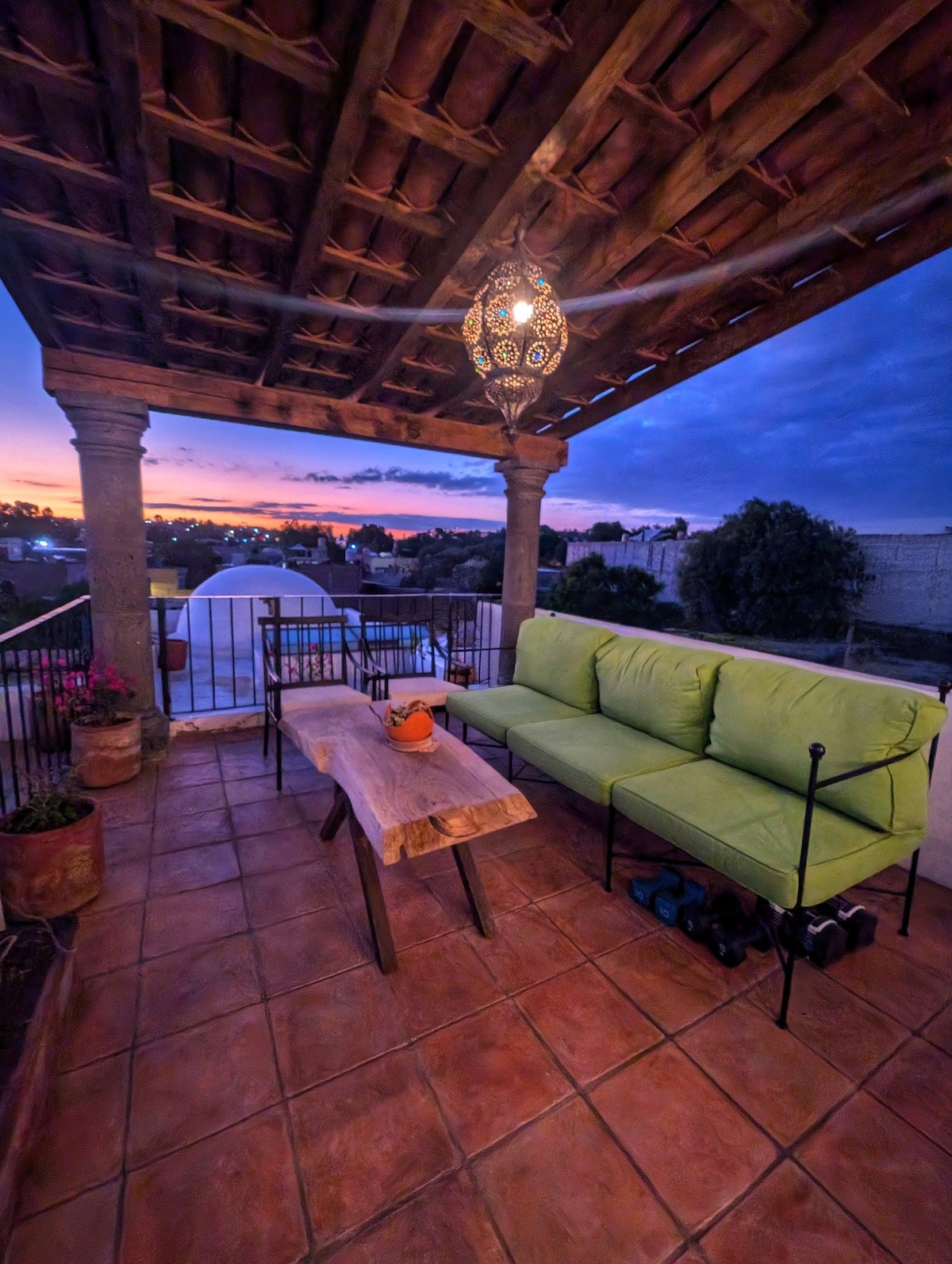 A covered rooftop terrace features vibrant green seating and a rustic wooden table. Potted plants line the railing, while a lantern overhead adds a soft glow. The sunset paints the sky with warm hues, enhancing the peaceful ambiance.