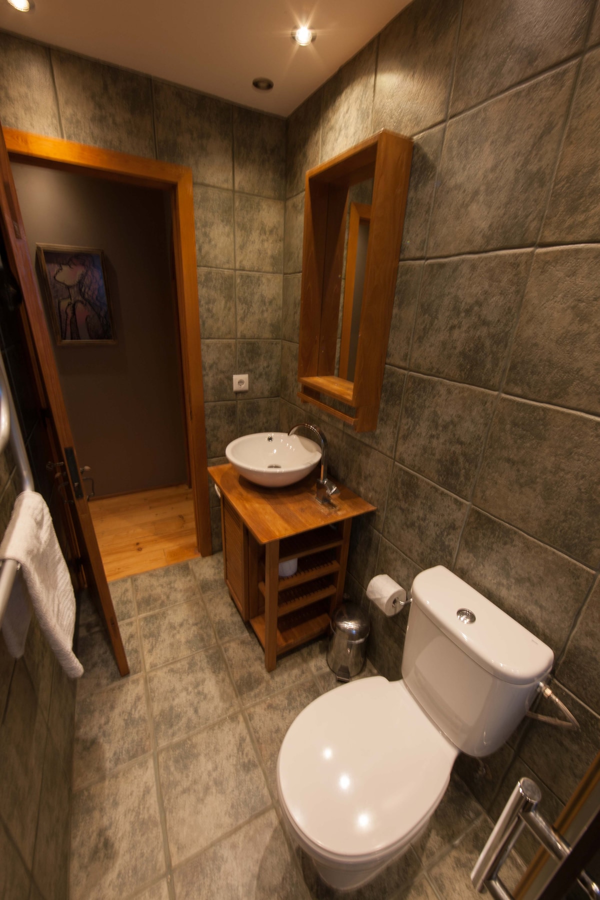 The bathroom features textured grey tiles, providing a modern feel. A wooden vanity with a round sink is positioned alongside a spacious mirror. A white toilet is present, along with a towel rack and wooden storage unit, enhancing the overall functionality of the space.