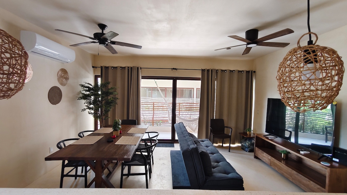 A bright living area features a comfortable sofa facing a wall-mounted television. A dining table is set with black chairs and a small centerpiece. Soft lighting is provided by woven pendant lamps, and large glass doors allow natural light to fill the space.