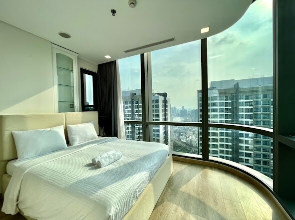 A modern bedroom features a spacious bed with crisp white linens, positioned against large windows offering a panoramic city view. The room is well-lit with natural light, highlighting the minimalist design and wooden flooring. Soft curtains frame the windows, adding an element of privacy.